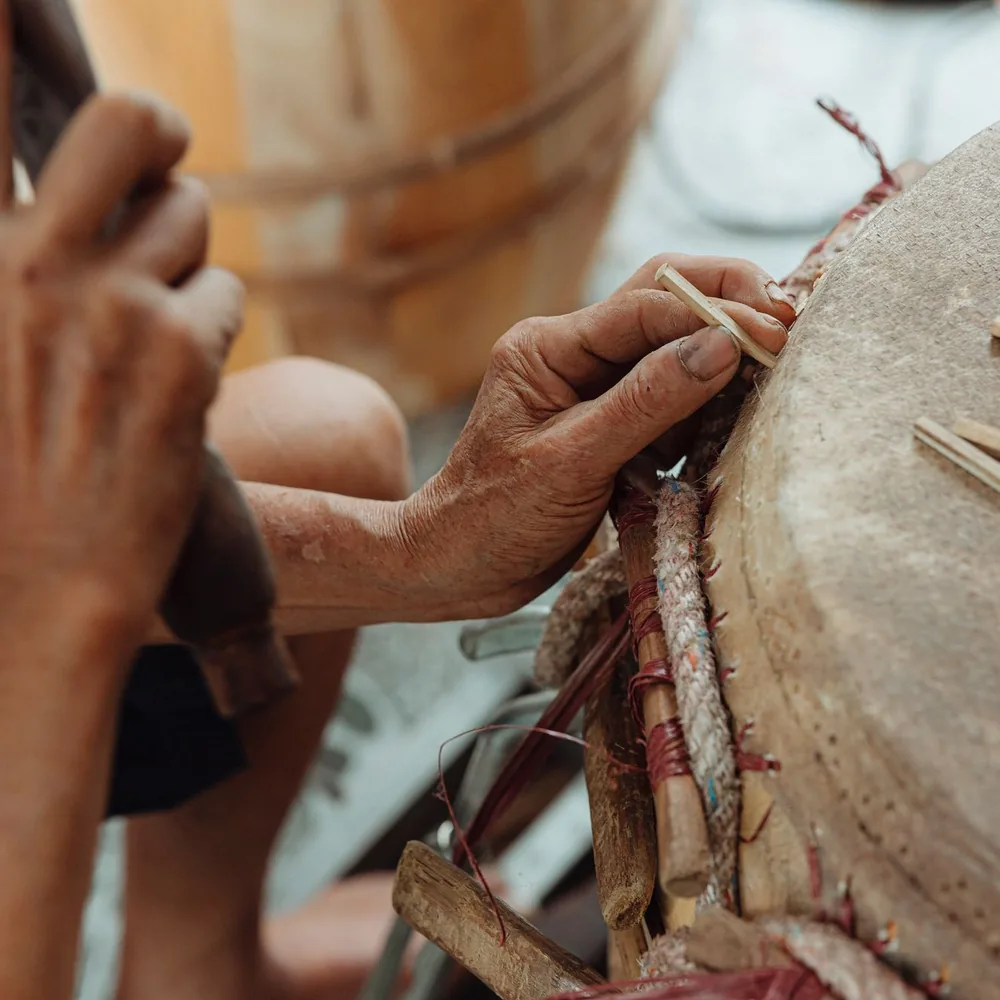 Manos trabajando un tambor de madera sobre un banco de taller, con herramientas alrededor